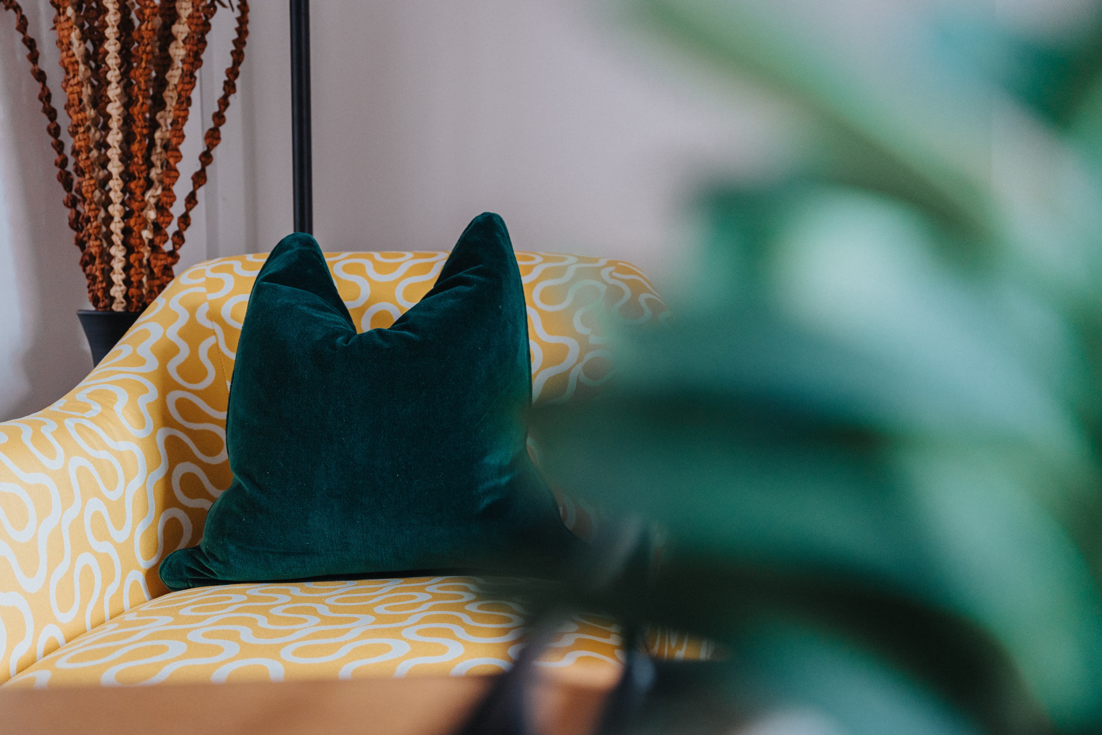 Dark green velvet pillow on a yellow and white patterned armchair behind blurry green leaves.